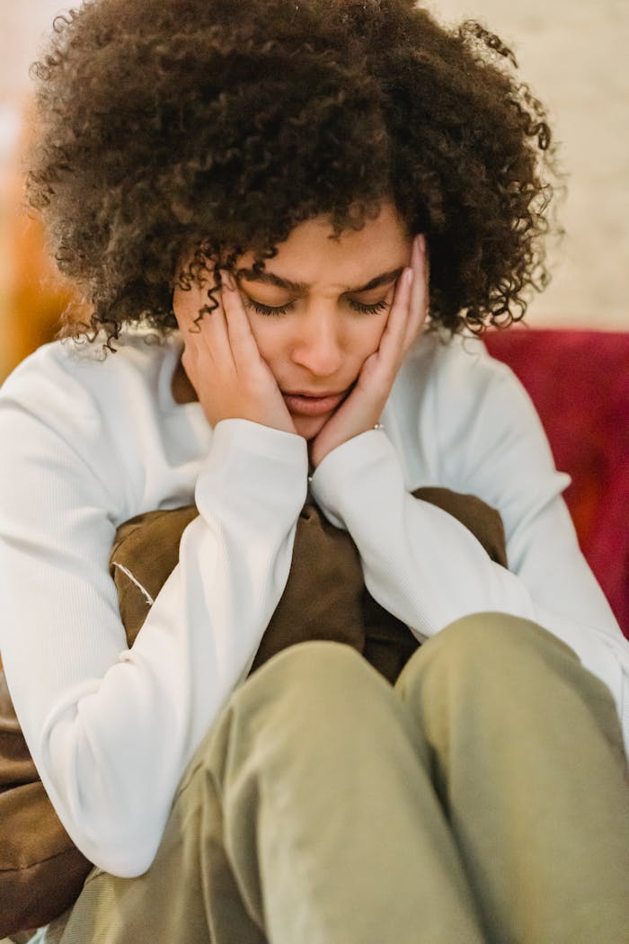 Young sad ethnic lady in casual outfit sitting with pillow with hands on head and closed eyes in light room