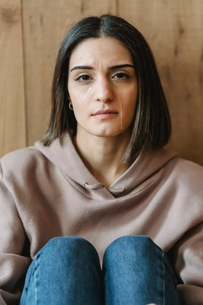 Young woman with tears looking serious, sitting indoors against wooden background.