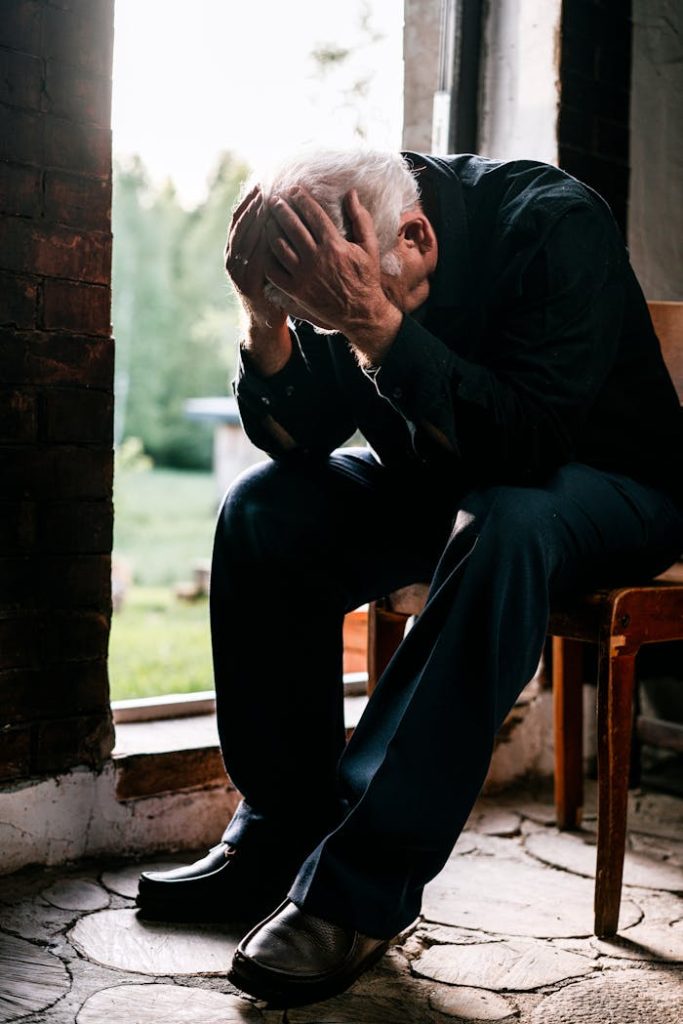 Senior man in black, grieving alone, sitting on a wooden chair by a doorway in a pensive pose.
